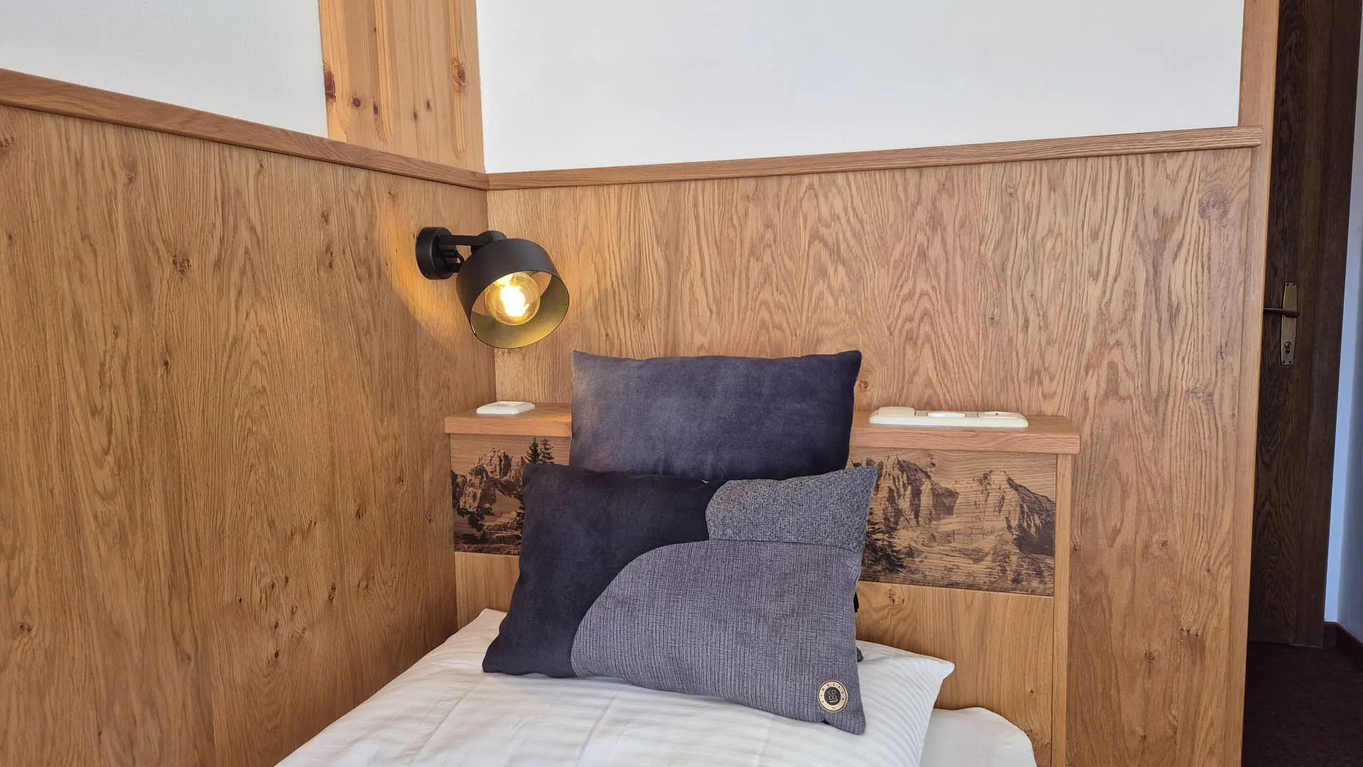 Holzwand mit Lampe und begrünten Kissen auf dem Bett